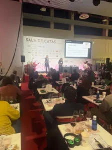 Sala de catas con pupitres individuales equipados con copas, orientados hacia un escenario con ponentes.